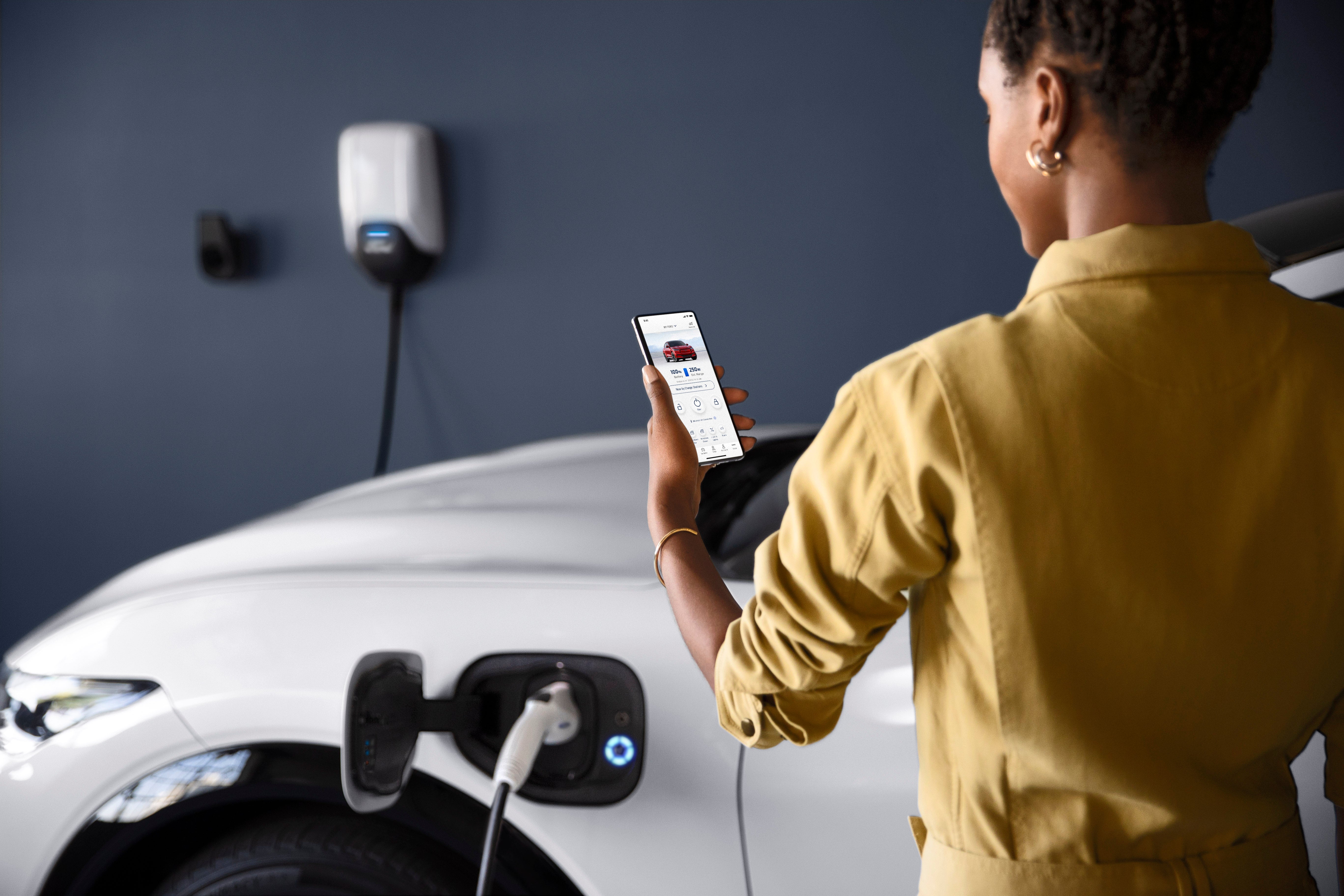 Woman standing beside a Ford Mach-E as she checks her phone and the vehicle charges, as it is all-electric